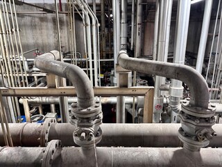 Heavy industrial piping network with large metal pipes, flanges, and valves running through a dimly...