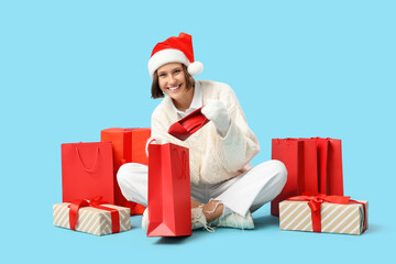 Beautiful young happy woman in Santa hat with shopping bags and gift boxes sitting on blue background. Christmas celebration