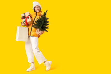 Beautiful young happy woman with shopping bags, gift box and Christmas tree on yellow background