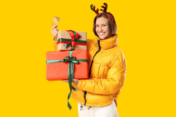 Beautiful young happy woman in reindeer horns with gift boxes and credit card on yellow background. Christmas celebration