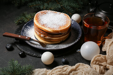 Plate of sweet pancakes with blueberries, Christmas balls and glass cup of tea on black background