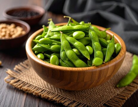 Rustic wooden bowl of fresh green edamame on woven mat with soybeans and dark sauce in background.