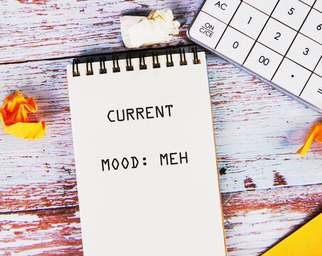 A notepad displays CURRENT MOOD MEH amidst crumpled paper and a calculator. The scene suggests a sense of indifference or boredom in a workspace setting