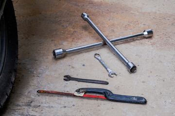 Automotive hand tools on concrete surface with visible tire, including lug wrench, hacksaw, and wrenches in realistic garage or workshop repair setup