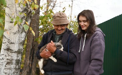 Elderly Russian grandfather and his adult granddaughter looking at a gray cat outdoors in village garden. Rural autumn family connection.