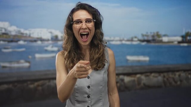 Young hispanic woman wearing glasses and a light grey vest points finger by sunlit seaside street; playful.