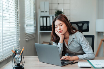Exhausted young Asian woman sleeping at office desk with laptop. Experiencing burnout, fatigue, and...