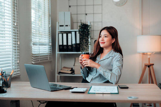 Young Asian business woman relaxing at office desk, holding coffee cup and looking away, taking a pleasant break from work