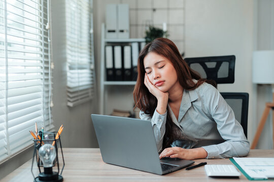 Exhausted young Asian woman sleeping at office desk with laptop. Experiencing burnout, fatigue, and lack of sleep - Powered by Adobe