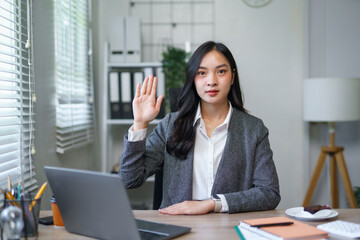 Young Asian woman raising hand during video call, making a sincere promise or voting in a corporate...