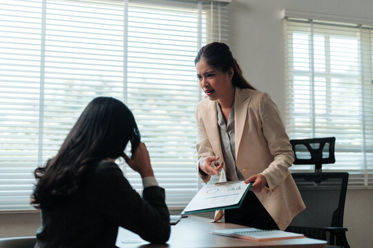 Angry female boss confronting a stressed employee, holding a document and discussing business problems in an office setting