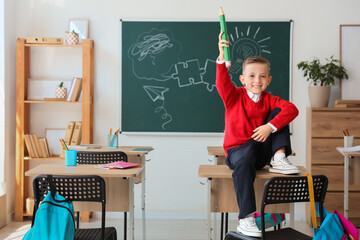 Cute little schoolboy with large pencil sitting on desk in classroom