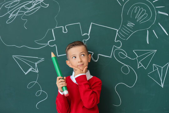 Thoughtful little schoolboy with large pencil near chalkboard in classroom - Powered by Adobe