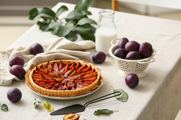Plate with tasty plum pie, colander, spatula and bottle of milk on table in kitchen