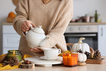 Young woman pouring tea from teapot into cup and candles burning on table in kitchen, closeup