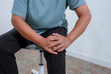 An elderly Asian woman sitting on a chair, gently holding her knee with both hands, expressing pain and discomfort caused by knee pain or osteoarthritis, reflecting aging joint problems.