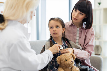 A doctor uses a stethoscope to examine a young girl, with the mother providing support. The scene takes place in a medical office.