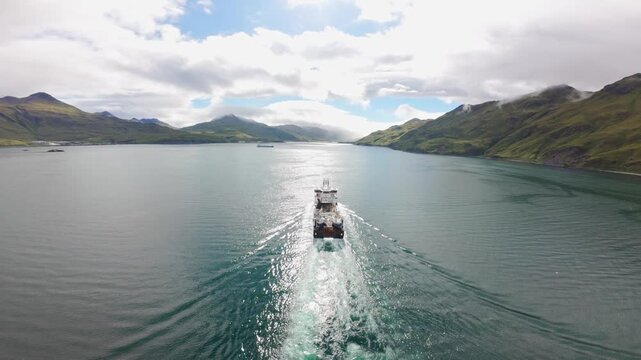 Aerial View Large Fishing Boat Moving Through Calm Alaska Waters. Commercial trawler sailing in Unalaska Bay, Alaska, with calm turquoise water and green mountains in the background.