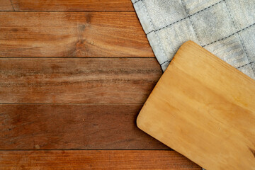 A top-down, flat lay view of a light-colored wooden cutting board overlapping a blue and white checked kitchen towel. They are set on a dark brown wooden plank surface, leaving clear copy space.