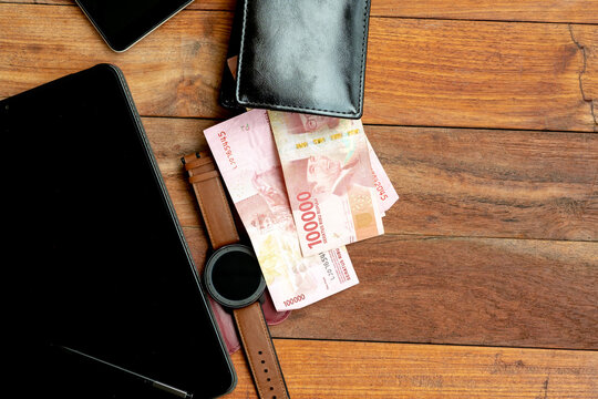 An overhead view of a dark wooden table with several one-hundred thousand Indonesian Rupiah banknotes, a black wallet, a black screen tablet, and a smartwatch.