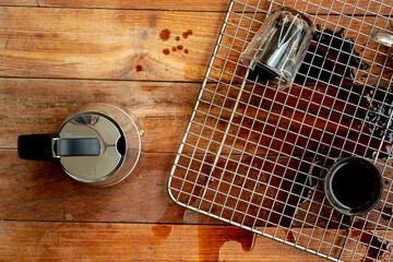 An overhead view of spilled black coffee on a rustic wooden table and a metal cooling rack. A coffee pot, a full glass, and an overturned glass are visible. © athian
