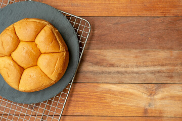 A top-down flat lay of a round, segmented bread roll on a dark slate plate. The plate sits on a wire cooling rack, all set against a warm, brown wooden table providing copy space.