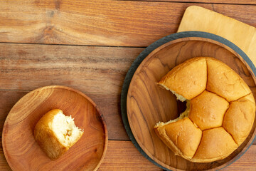 A top-down flat lay of a torn, segmented bread roll. A single piece is on a small wooden plate, while the main loaf sits on a larger wooden plate, all on a wood table.