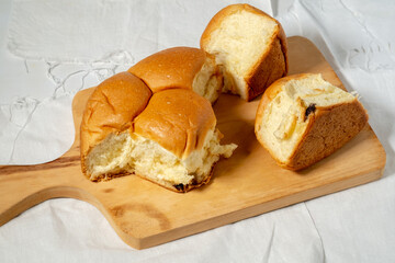 A close-up of a soft, golden-brown bread roll torn into several pieces, revealing the fluffy interior, all placed on a wooden cutting board over a white cloth.