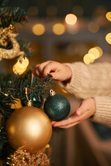 Child decorating Christmas tree, holding gold and green ornaments with both hands, focusing on placing bauble on branch, festive lights softly glowing in background