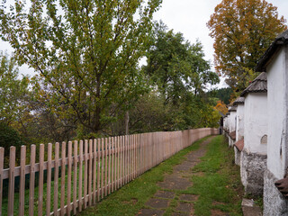 Fototapeta premium Old churchyard wall and wooden fence along narrow path in Špania Dolina, Slovakia, surrounded by trees with autumn leaves in a peaceful mountain village.