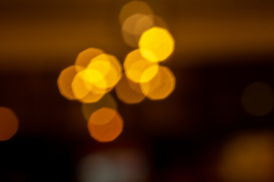 An abstract photograph of warm, golden-yellow bokeh lights. The out-of-focus light spots are clustered together, revealing a faint octagonal shape against a dark, blurred background.