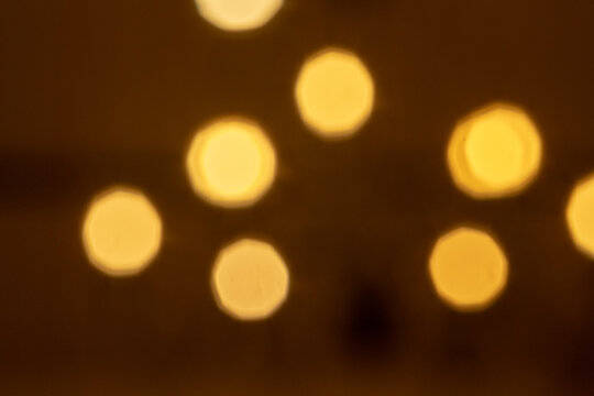 An abstract photograph of warm, golden-yellow bokeh lights. The out-of-focus spots are scattered against a dark, warm-toned brown background, showing a faint octagonal shape.