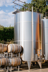 Modern stainless steel wine fermentation tank and traditional oak barrels in an outdoor winery setup under natural daylight