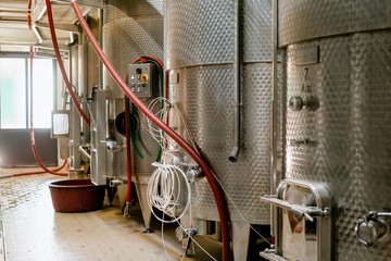 Interior of a modern winery with stainless steel fermentation tanks, control panels, and industrial hoses for wine production process