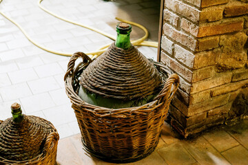 Vintage green glass demijohn bottles with woven wicker covers and handles, placed on tiled floor near brick wall, traditional wine storage container
