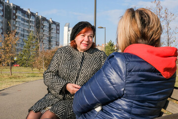 Two middle-aged women are talking while sitting on a bench in a city park on a sunny autumn day