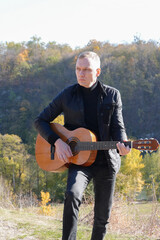Portrait of smiling caucasian artist plays wooden acoustic guitar in autumnal park. Young songwriter plays string musical instrument hobby outside in nature fall time. Audio music healing in natural