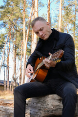 Beautiful blonde man playing acoustic guitar and singing relaxing enjoying outdoors on sunny autumn day. Artist learning and practicing playing musical instrument. Fall leaves autumnal weather 