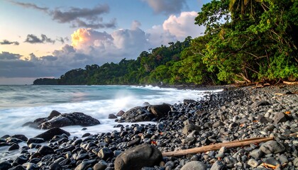 Beach rocks and lush vegetation line a coastline under a clouded sky at dusk, waves gently crashing
