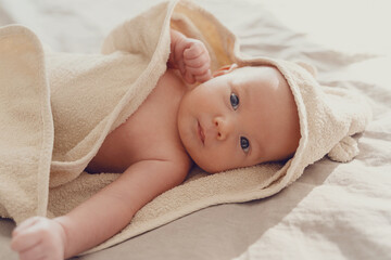 Happy curious baby child after bath with towel on head. Adorable little boy wrapped in blanket. Cutest 3 months old baby smiling in towel on bed close up at home. Child caring routine.