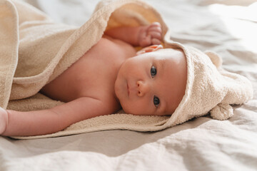 Happy curious baby child after bath with towel on head. Adorable little boy wrapped in blanket. Cutest 3 months old baby smiling in towel on bed close up at home. Child caring routine.