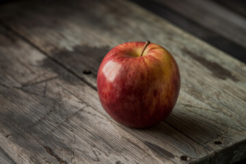A single, crisp red apple with yellow streaks sits on a rustic, weathered wooden surface, illuminated by a gentle, soft light.