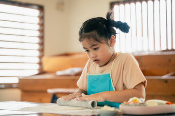 Young girl engaged in playful cooking activity, rolling dough on table, focused expression,...