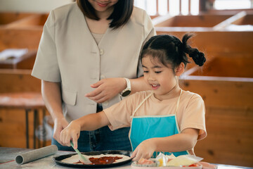 Mother and daughter enjoy cooking together in kitchen, child learns to spread sauce on pizza base,...