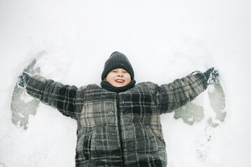 Caucasian child lying on snow making snow angel, smiling with arms outstretched, wearing winter...