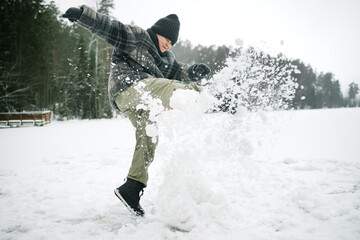 Caucasian teenage boy kicking snow outdoors, wearing winter hat and gloves, standing on snowy field...