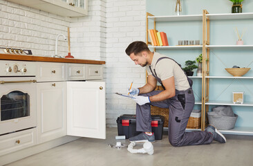 Experienced plumber in workwear crouching on kitchen floor and repairing sink pipes under cabinet. Male technician using tools to remove blockage and restore proper water flow during home visit.