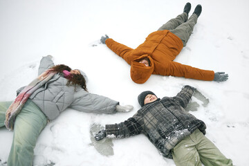 Three people lying on snow making snow angels, including Caucasian man, woman, and child, all...