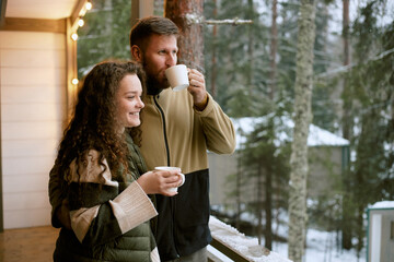 Caucasian young woman and man standing on balcony drinking hot beverages together, smiling and looking at snowy forest landscape during winter morning
