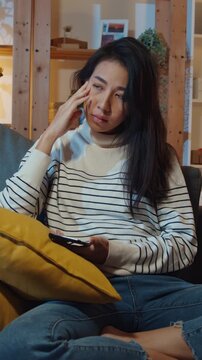 Vertical Screen : Thoughtful Asia lady holding phone feeling sad waiting for call sit at sofa in living room at house night feel lonely, Sad depressed teenager spend time alone, Social distance.